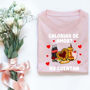 May include: Pink t-shirt with a white graphic that says "Calorias de Amor? No Cuentan" and features a cartoon image of Mexican food, drinks, and hearts.