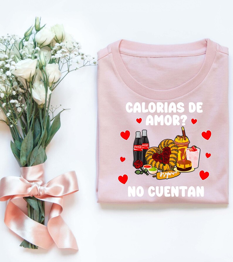 May include: Pink t-shirt with a white graphic that says "Calorias de Amor? No Cuentan" and features a cartoon image of Mexican food, drinks, and hearts.