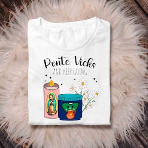 May include: White t-shirt with the text "Ponte Vicks and Keep Going" in black. The design features a pink candle with a depiction of the Virgin Mary, a blue Vicks VapoRub container with a cartoon face, and white daisies.