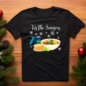 May include: Black t-shirt with the text "Tis the Season" in white, featuring a bowl of soup with vegetables, a container of Vicks VapoRub, and tortillas. Snowflakes are scattered around the design.