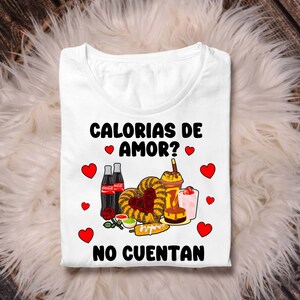 May include: White t-shirt with a graphic design featuring a variety of Mexican foods, including churros, corn on the cob, and Coca-Cola. The text on the shirt reads "Calorias de Amor? No Cuentan".