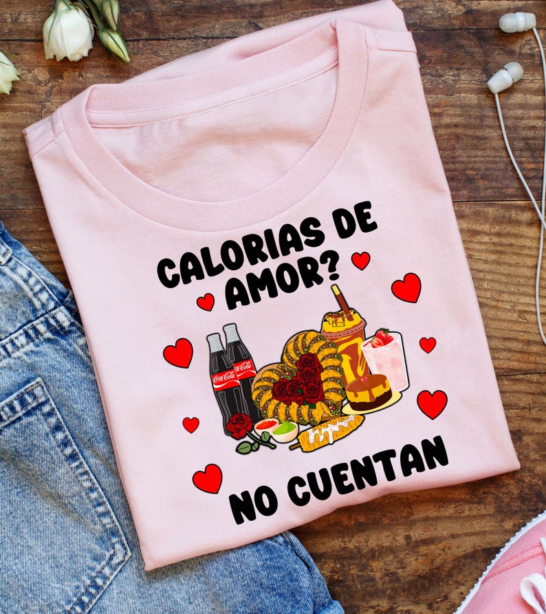 May include: Pink t-shirt with the text "Calorias de Amor? No Cuentan" in black. The shirt features a graphic of various Mexican foods, including churros, a heart-shaped pastry, and a corn dog, with red hearts scattered around the design.