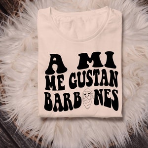 Op de afbeelding: Een beige T-shirt met zwarte tekst die "A mi me gustan barbones" luidt met een cartoon-kerstmangezicht in het midden van de tekst.