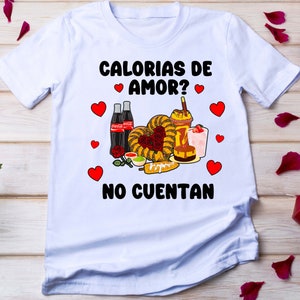 May include: White t-shirt with a graphic design featuring the text "Calorias de amor? No cuentan" in black. The graphic also includes a heart shape made of various Mexican foods, including churros, corn on the cob, and a glass of horchata.