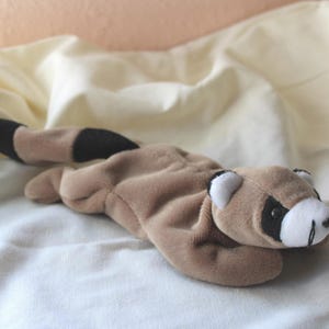Può includere: Un peluche a forma di procione nei toni del marrone, beige, nero e bianco, appoggiato su una superficie di tessuto bianco. Il procione ha una maschera nera intorno agli occhi, una coda a strisce nere e beige e dettagli bianchi sulle orecchie e sul viso.