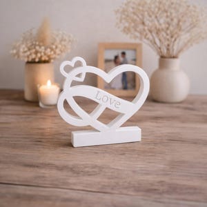 Peut inclure: Décoration blanche en forme de cœur en bois avec le mot "Love" gravé. Le design comprend un symbole infini et un petit cœur. La décoration est posée sur une surface en bois, avec une bougie et des fleurs en arrière-plan.