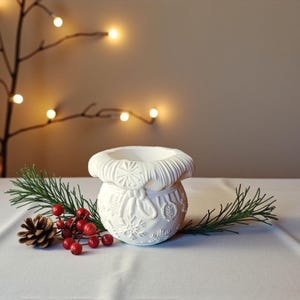 Puede incluir: Un portavelas de cerámica blanca con un diseño en relieve de copos de nieve y adornos. El portavelas está decorado con una rama de pino, bayas rojas y una piña. El fondo presenta una cadena de luces.