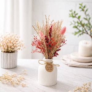 Könnte beinhalten: Eine weiße, gerippte Keramikvase enthält einen Strauß getrockneter Blumen in Rosa-, Rot- und Beigetönen. Ein kleines, rundes Etikett mit der Aufschrift "Handmade with love" ist mit Bindfaden befestigt. Eine weitere Vase mit getrockneten Blumen befindet sich im Hintergrund.