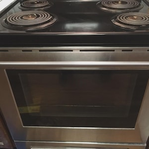 May include: Stainless steel electric range with a black cooktop and four coil burners. The oven door has a large glass window and a stainless steel handle. The range is set into a wooden cabinet.