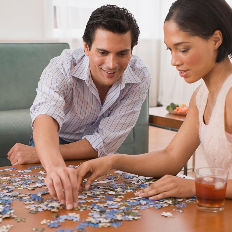 Pode incluir: Um casal montando um quebra-cabe&ccedil;a em uma mesa marrom. As pe&ccedil;as do quebra-cabe&ccedil;a s&atilde;o principalmente azuis e brancas. Um copo de l&iacute;quido &acirc;mbar est&aacute; sobre a mesa. O homem usa uma camisa listrada e a mulher uma blusa sem mangas.