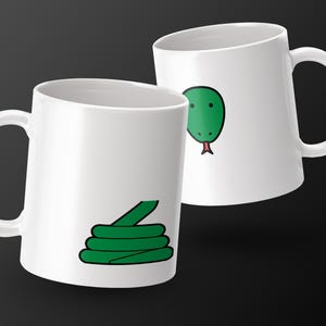 May include: Two white ceramic mugs with a green snake design. One mug features a coiled snake, while the other displays a snake head with a red tongue. The mugs have a glossy finish and a curved handle.