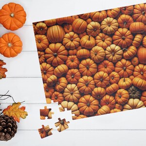 May include: A jigsaw puzzle with an autumnal theme, showing a close-up of numerous orange pumpkins. The puzzle is partly assembled, with a few pieces separate. Two small pumpkins, a pinecone, and autumn leaves are on a white wooden surface.