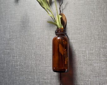 May include: A brown glass bottle with a metal wire stem attached to a wooden wall mount. The bottle is filled with white flowers and green leaves.