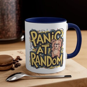 May include: White ceramic mug with a blue rim and handle. The mug features a cartoon bear with the text "Panic at Random" in yellow, blue, and red.