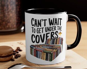 Puede incluir: Taza de cerámica blanca con borde y asa negros. La taza presenta una ilustración en blanco y negro de una cama hecha de libros con una lámpara en la parte superior. El texto "Can't wait to get under the covers" está impreso en la taza.