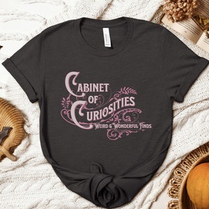 May include: A dark grey t-shirt with a pink graphic that says "Cabinet of Curiosities Weird & Wonderful Finds".