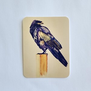 May include: A rectangular card featuring a watercolor illustration of a raven perched on a wooden post. The raven is depicted in shades of black, purple, and gold. The post is a light brown color, and the background is a soft beige.