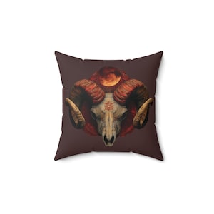 May include: Square throw pillow with a dark brown background featuring a detailed illustration of a ram skull with large, curved horns. The skull is cream-colored, with a red and orange halo and a celestial design.