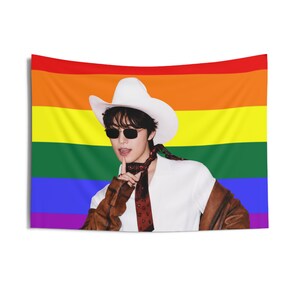 Pode incluir: Uma tapeçaria retangular com uma bandeira arco-íris horizontal ao fundo. Em primeiro plano, uma pessoa usa um chapéu de cowboy branco, óculos de sol escuros, uma camisa branca, um lenço escuro estampado e uma jaqueta de cor castanha.