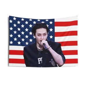 May include: A wall tapestry featuring a person holding a microphone in front of an American flag. The person is wearing a black jacket and a silver chain. The flag has blue stars and red and white stripes.