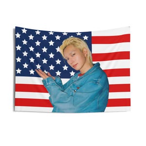 May include: A wall tapestry featuring the American flag with red and white stripes and a blue field of white stars. A person with blonde hair and a denim jacket is in front of the flag.