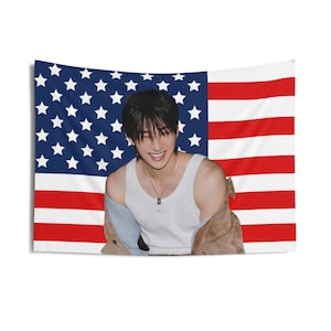 May include: A wall tapestry featuring the American flag as a backdrop. A person is wearing a white tank top and a brown jacket. The person is smiling and looking at the camera.