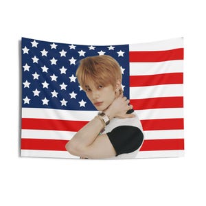 May include: A rectangular wall tapestry featuring the American flag in red, white, and blue. A person is positioned in front of the flag, wearing a black and white shirt and gold jewelry. The tapestry is likely made of fabric.