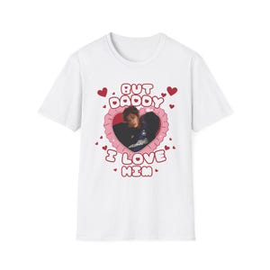 May include: White t-shirt featuring a heart-shaped design with the text "BUT DADDY I LOVE HIM" in red. The heart contains a photo of a person. Small red hearts are scattered around the main design.