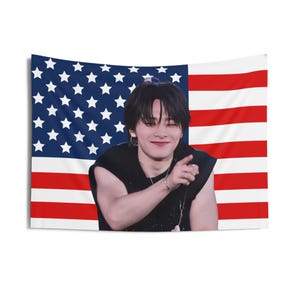 May include: A tapestry featuring the American flag as a backdrop. In front of the flag is a person wearing a black sleeveless top, smiling and pointing. The flag has red and white stripes and a blue field with white stars.