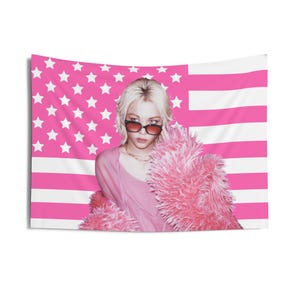 以下が含まれることがあります： サングラスとピンクの衣装を着た金髪の人物と、ふわふわのピンクのジャケットが特徴のピンクのタペストリー。背景は、白い星とストライプが入ったピンクのアメリカ国旗です。