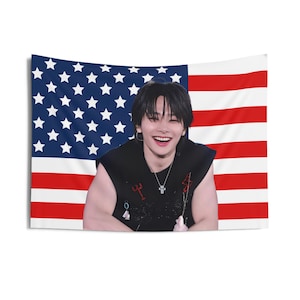May include: A rectangular wall tapestry featuring a smiling person in front of an American flag. The flag has red and white stripes and a blue field with white stars. The person is wearing a black sleeveless top and a silver cross necklace.