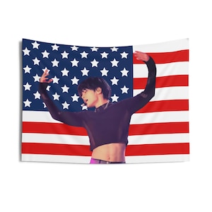 May include: A tapestry featuring the American flag as a backdrop. In front of the flag, a person is wearing a black long-sleeved crop top and purple pants. The person is in a dance pose with arms raised.