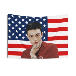 May include: A tapestry featuring the American flag as a backdrop. In front of the flag is a person wearing a red shirt and a silver watch. The person is touching their face with their hand. The tapestry is rectangular and hangs on a white wall.