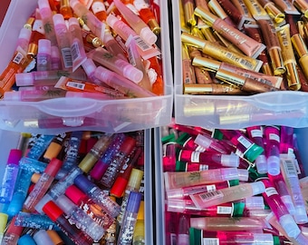 May include: A collection of lip gloss tubes in clear plastic containers. The glosses are in various colors, including pink, red, gold, and clear, with different applicators and packaging. The containers are filled with a variety of lip gloss products.