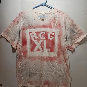 Peut inclure: T-shirt blanc avec un effet de peinture en aérosol rouge et noir. Le t-shirt présente un carré rouge avec les lettres "RCG" au-dessus de "XL" en blanc. Il est suspendu à un cintre bleu.