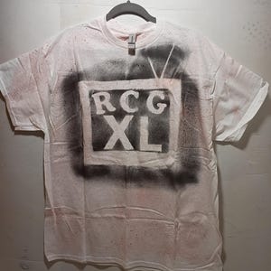 May include: White t-shirt with a black and gray spray-painted television design. The design includes the letters "RCG" above "XL" in a rectangular frame. The shirt has a crew neck and short sleeves.