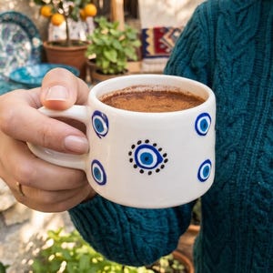 May include: A white ceramic mug featuring a hand-painted blue and black evil eye pattern, filled with a dark brown beverage topped with cinnamon. The mug has a rounded handle and a smooth finish, showcasing a repeating circular eye motif.