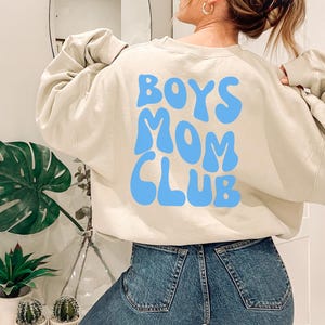 Könnte beinhalten: Ein hellbeiger Pullover mit dem Text "Boys Mom Club" in blauer Schrift. Die Buchstaben sind in einer Retro-Wellenschrift.