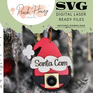 May include: A wooden Christmas ornament shaped like Santa Claus with a camera lens in place of his eye. The ornament is painted red and black and has the words "Santa Cam" written on it.