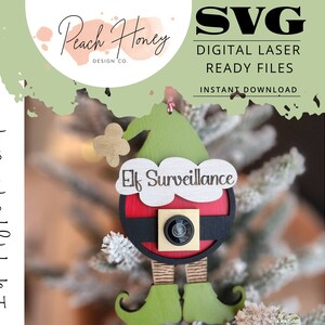 May include: A green elf ornament with a red and black Santa suit and a camera lens. The ornament has the text "Elf Surveillance" on it.