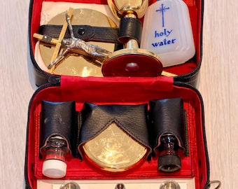 Puede incluir: Un estuche negro con forro rojo que contiene objetos religiosos. Incluye un cáliz dorado, un crucifijo, una botella con la etiqueta "holy water" y otros accesorios litúrgicos. El estuche está abierto, revelando el contenido.