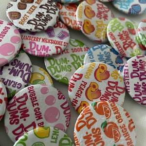 May include: A collection of colorful Dum Dums lollipops, each wrapped with a different flavor label. Flavors include Root Beer, Bubble Gum, Orange, and Strawberry Milkshake. The candies are round and have a white background with the flavor name in a bold font.