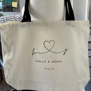 May include: A white canvas tote bag with black handles. The bag has a black heart design with the initials "H" and "J" intertwined. The text "HOLLY & JASON" and the date "04-24-24" are printed below the heart.