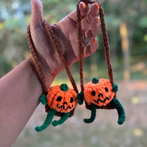 May include: Two orange crochet pumpkin ornaments with black facial features and green stems and legs. The pumpkins are suspended by brown cords, perfect for Halloween decorations.