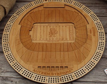May include: A circular wooden cribbage board featuring a laser-engraved replica of Autzen Stadium. The centre field displays the Oregon logo and text, while the perimeter includes drilled holes for scoring. The board is light brown with dark wood grain.