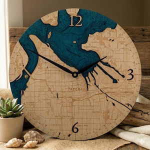 May include: A round wooden clock with a map design. The clock face features a blue water area and the city of Tacoma, with black hands indicating the time. The clock is approximately 12 inches in diameter.