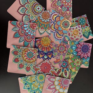 May include: A selection of pink pouches, each adorned with a detailed mandala design. The pouches display a variety of colourful bead patterns in blue, yellow, red, and green, forming a mosaic. The pouches are arranged on a dark surface.