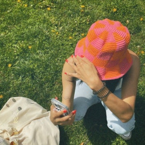 May include: A person wearing a pink and orange crocheted bucket hat, blue jeans, and a white tote bag. The person is holding a clear phone case with a white phone inside.