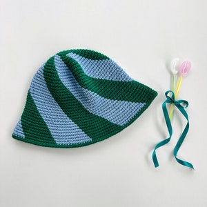 Puede incluir: Un sombrero de sol de crochet a rayas verdes y azules. El sombrero está hecho de un hilo suave y texturizado y tiene un ala ancha.