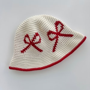 Puede incluir: Un sombrero de sol blanco de ganchillo con un borde de ganchillo rojo y diseños de lazo rojo.